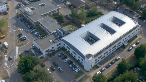 An aerial view of the large Derwent Centre complex in Harlow. It features a modern, white, curved-roof structure at the centre. Surrounding it are several older, rectangular buildings arranged along internal roads and parking areas. Cars are parked in multiple spaces and the site is bordered by trees.