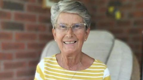 Susan Whittles - a woman with short grey hair and clear glasses is smiling into the camera and sitting in a chair. She is wearing a yellow and white-striped top and necklace with white-stud earrings. The background is blurred but you can make out a brick wall behind her.
