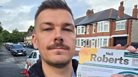 A picture of Neil Roberts stood in a Cardiff Road surrounded by terraced houses and cars in the background. He is holding a set of leaflets emblazoned with his own name and the Plaid Cymru yellow poppy logo.