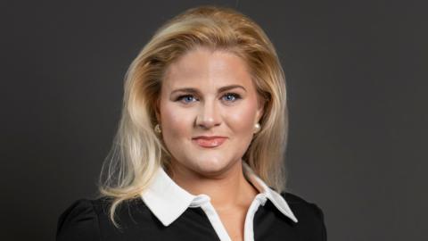Alex Davies-Jones is smiling in the head and shoulders picture. She has-shoulder length blonde hair which falls on a black top with a large white collar. She is wearing large pearl earrings.