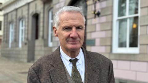 Deputy Tom Binet pictured in the Royal Square with the States building visible behind him. He's wearing a brown suit, white shirt and green tie. He has short white hair and is smiling.