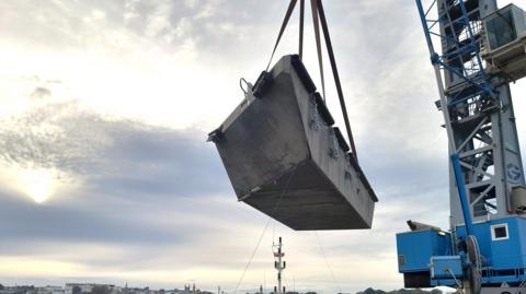 A picture of the berth being lifted off the ground by a crane. It is a grey coloured block with a blue coloured crane. In the distance there is a blue sky with clouds and properties in the distance.