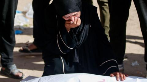 A crying woman wearing dark clothing and a hijab kneels, one hand to her mouth, the other on the shrouded body of a corpse 