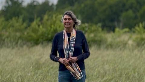 Helen Lewy is pictured standing in a field looking up to the sky. She is an older woman with dark grey hair that is blowing in the wind. Helen is wearing a navy jumper and a navy, pink, yellow and cream patterned scarf, the bottom of which she is holding.