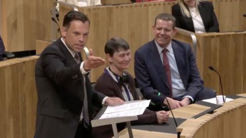 A man in a black suit is gesturing towards someone off screen in the Senedd, while a woman in a brown cardigan and a man in a blue suit with red tie are in the background smiling. There are also wooden seating areas and wired microphones coming out of the in built wooden desks within the Senedd. 