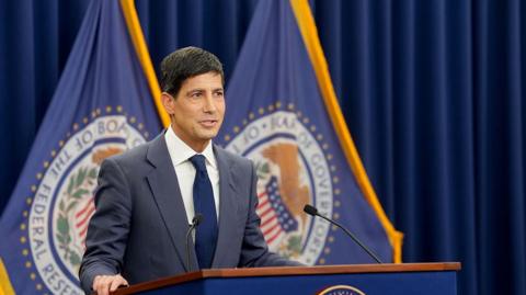 Kevin Warsh stands at a podium giving a press conference, wearing a navy suit, navy tie and white shirt, flanked by flags with the crest of the Federal Reserve.