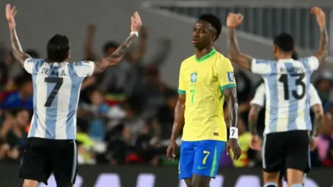 Vinicius Jr looking sad with two celebrating Argentina players in the background.