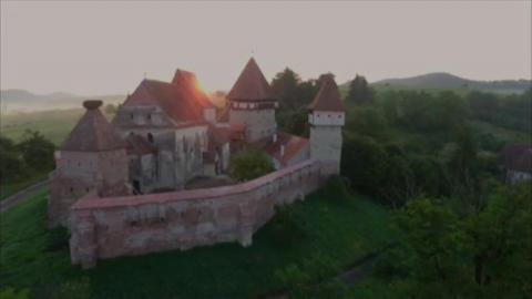 A castle and walls sitting on a green hill, surrounded by green space. The sun is rising in the background