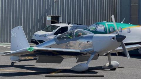 Matthews inside his silver Vans RV‑7. It is a small aircraft parked on a tarmac runway.