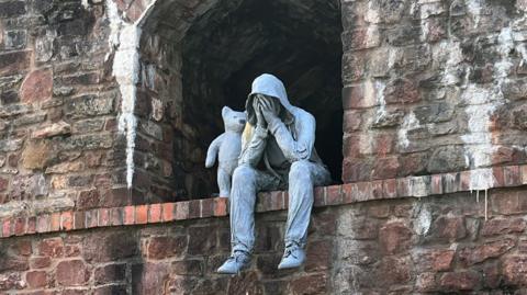 A grey statue of a person wearing trainers, trousers and a hoodie. They are sitting on the ledge of a brick archway with their head in their hands and their hood up. A small bear stands beside them, with one paw resting on the person's shoulder. 