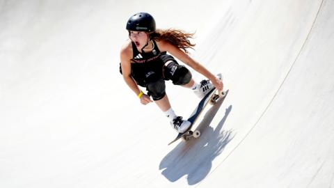 Lola Tambling skateboarding - a girl with long brown hair wearing a black helmet with her mouth wide open, wearing a black vest saying GREAT BRITAIN on it, wearing white trainers with three stripes on them, black laces, black knee pads, holding one hand on her skateboard, she is going downhill in a skateboard bowl which is white