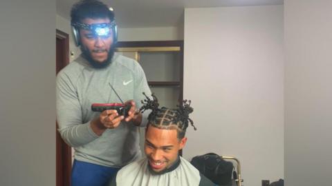 A man wearing a head torch trims another man's hair with clippers in a room with a wooden cupboard and metal-framed chair visible in the background.