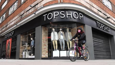 A woman in a mask cycles past a closed Topshop store