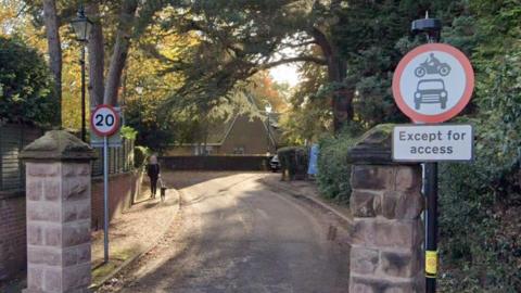 A winding narrow road with a sign at the entrance saying no entry to cars and motorbikes except for access and trees lining both sides