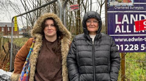 Bethany Wood, with dark hair, wearing a fur-lined hooded coat, standing next to Helen Fisher, who is wearing glasses and has a black puffer coat on with the hood up. Behind them is a metal fence with a sign that reads land for sale, sold. There is overgrown grass and bushes behind the fence. It is a wet day. 