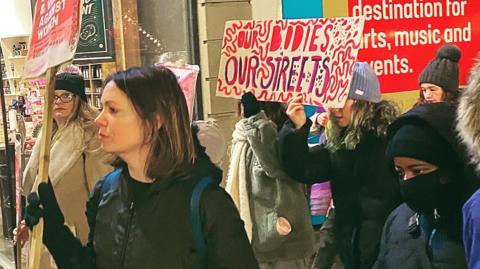 A number of women of various ages, wearing warm clothing and carrying banners as they walk through Sheffield city centre on a Reclaim the Night march in 2022. One of the banners reads "Our bodies, our streets".