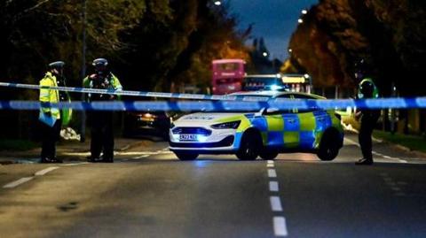 A police car is parked diagonally across the road which is taped off. Three officers are standing in the road. It is dark and the scene is lit by street lamps.