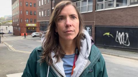 A young woman with shoulder length brunette hair looks at the camera. She's wearing a number of layers including a green winter coat and grey hoodie.