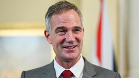 Peter smiles while standing in front of a blurry pale yellow background. He is wearing a light grey suit jacket, white shirt and deep red tie.