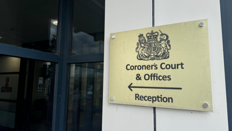 An entrance to a coroner's court with a close up of a gold sign with black writing that reads: "Coroner's Court & Officers", with an arrow pointing to a glass door. 