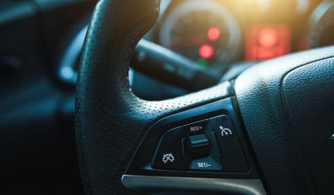 A close-up of a car steering wheel, with controls in shot and a blurred background featuring a speedometer on the vehicle's display panel.