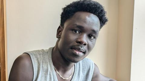 A young man with dark skin wearing a gold chain and grey vest top in a room with beige walls.