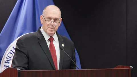 Tom Homan in a dark suit behind a wooden lectern