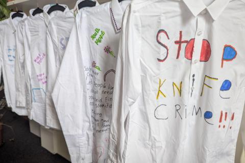 White long-sleeved school shirts hanging on a rack from black hangers depicting colourful artwork related to bringing an end to knife crime. The first of these is in red, blue, yellow and black with the words "Stop Knife Crime" on the front on the shirt.