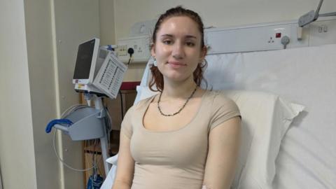 A young woman with dark hair sits upright in a hospital bed. She has a cannula in her arm.