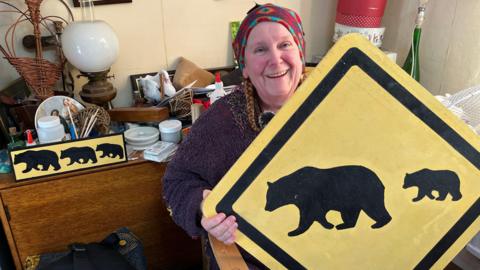 A woman wearing a brightly-coloured knitted headpiece and a purple jumper smiles as she sits on a chair with many different items on top of cabinets behind her. She is holding a large, yellow, diamond shaped sign with two black bear symbols on it. There is another, smaller, rectangular sign with three bears on it on the table behind her.