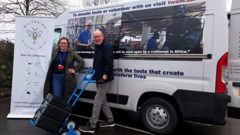 A man and a woman pose in front of a Wiltshire Digital Drive minibus and banner, as the man pushes a trolley with laptops on it.