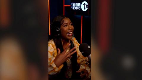 A woman in a radio studio smiles with one hand raised. 