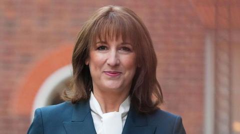 Rachel reeves who has bobbed brown hair and is wearing a white blouse and navy blazer, smiling.