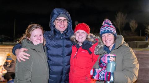 Four people standing next to each other all of them are wearing thick winter jackets and they all have a smile on their faces looking at the camera, with the second person to the left putting his arms around the group. 