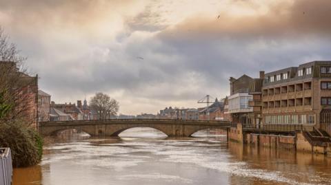A 19th century stone bridge spanning a river swollen with flood water. There are buildings on each side and a cloudy sky above.