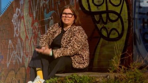 A woman in a leopard print jacket sits infront of a wall with graffiti on it