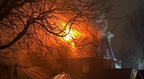 Large brick building with conservatory to its side - it is night and flames are seen coming through the roof of the building.