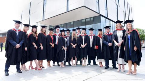 There are 16 people wearing caps and gowns smiling and standing outside a university building