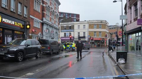 Lewisham High Street - a road lined with shops on both sides - closed, with police tape running across it and a police officer standing in the road.