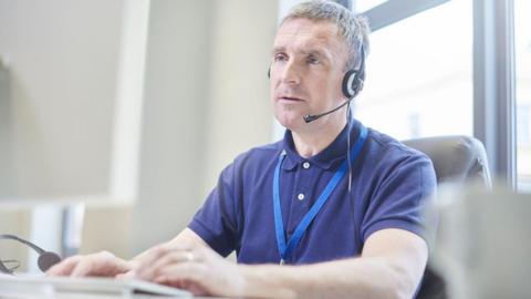 A stock image of a man on a video call. He is sitting at a desk wearing a headset and looking at a monitor.