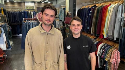 Harrison Farrell-Forster, right, is wearing a black T-shirt and is standing next to Dan, who is wearing a beige coloured jacket. They are standing in front of various clothing rails which have been prepared for the opening day of Wear. 