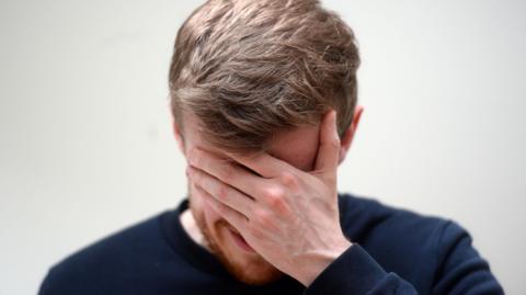 A man holds his face in his hand. He is looking down and wearing a blue top.