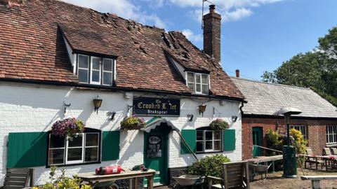 A view of the Crooked Billet Stoke Row on a sunny day. The damage to the roof following the fire is visible. The area around the building is taped.