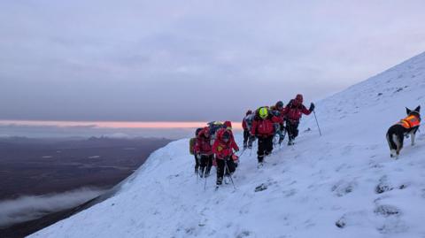 A group of people in red coats and mountain gear are walking on the slope of a mountain that is covered in white snow. There is a dog in an orange vest with them. They are dragging a man in a red stretched and have walking poles. In the distance you can see land all around and clouds below.