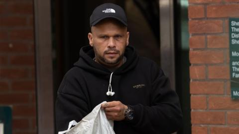 A man with a goatee is dressed in black and wearing a cap while holding a plastic bag.