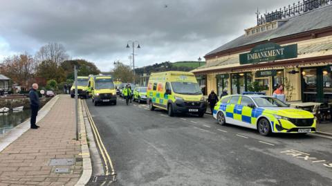 Multiple police and ambulance vehicles are parked on the road near the river. The Embankment Bistro on the right hand side. A man, wearing black, is stood watching the emergency services.