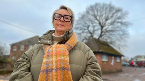 Councillor Denise Burke stood in a car park with a large oak tree in the background. She is wearing a green coat with an orange scarf, and has dark rimmed spectacles on her face.