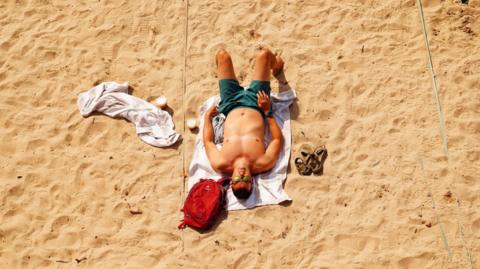 A man sunbathes on a sandy beach. The man is wearing green swimming shorts and sunglasses and he is laying on a towel. There is another towel beside him as well as a small red rucksack and a pair of brown sandals.
