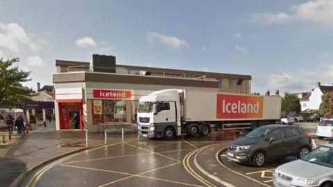 A front view of Iceland in Warminster under a blue sky, some of a car park to the right and pedestrians to the left. A large Iceland truck is in front.