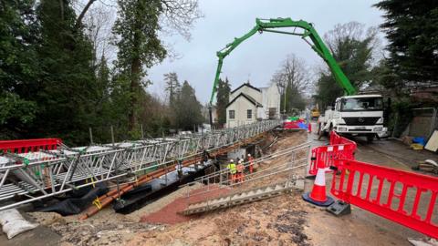 Construction work on a road with a temporary metal bridge structure in place. Workers in high-visibility clothing are seen beneath the bridge, while a large green concrete pump extends from a white lorry on the right. Red safety barriers and traffic cones line the site, with trees and a white house visible in the background under an overcast sky.